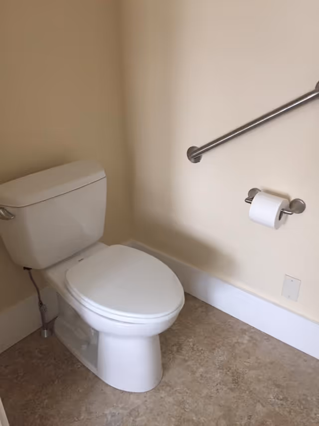 White toilet beside a beige wall with a metal grab bar and a toilet paper holder.