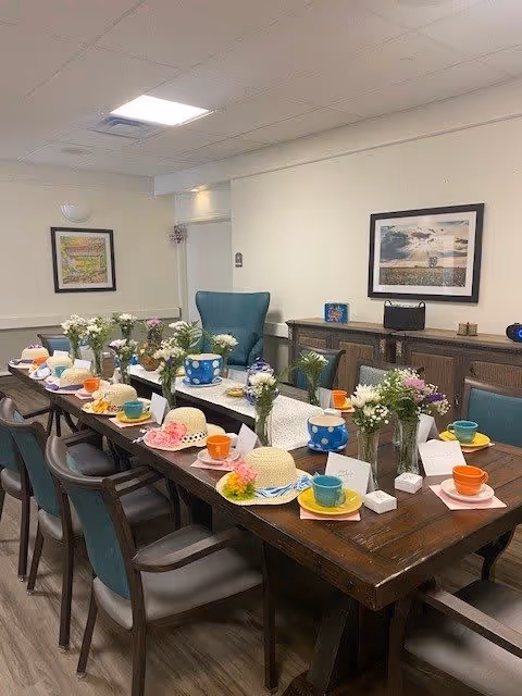 A long wooden dining table set for a gathering with colorful cups and saucers, floral centerpieces in glass vases, and decorative straw hats with flowers placed at each setting. The room has teal cushioned chairs around the table, framed artwork on the walls, and a wooden sideboard against the far wall.