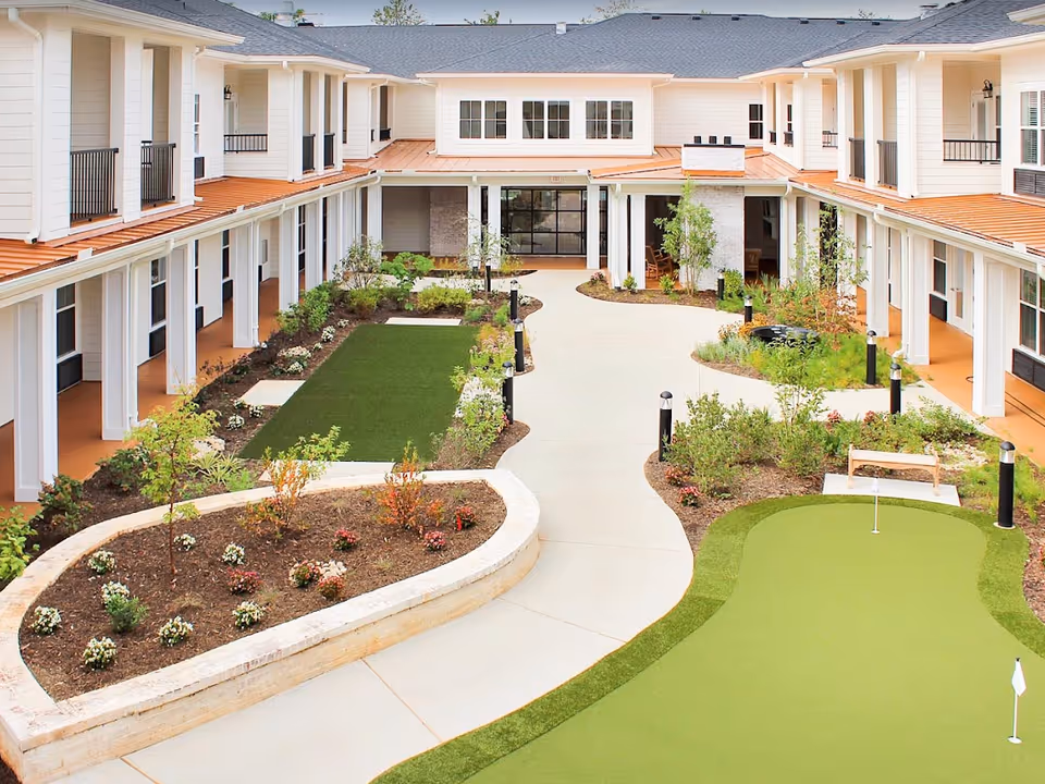 Landscaped central courtyard of a senior living facility with walking paths, raised planters, and a small putting green surrounded by a two-story building.