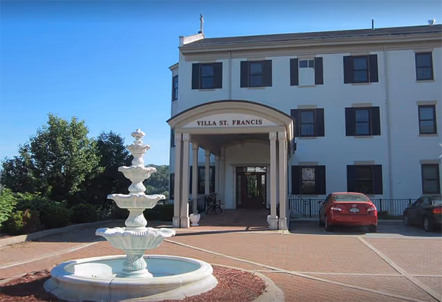 Front exterior view of Villa St. Francis building with a covered entrance supported by columns, a three-tiered white fountain in the foreground, and two parked cars on the right side under a clear blue sky.