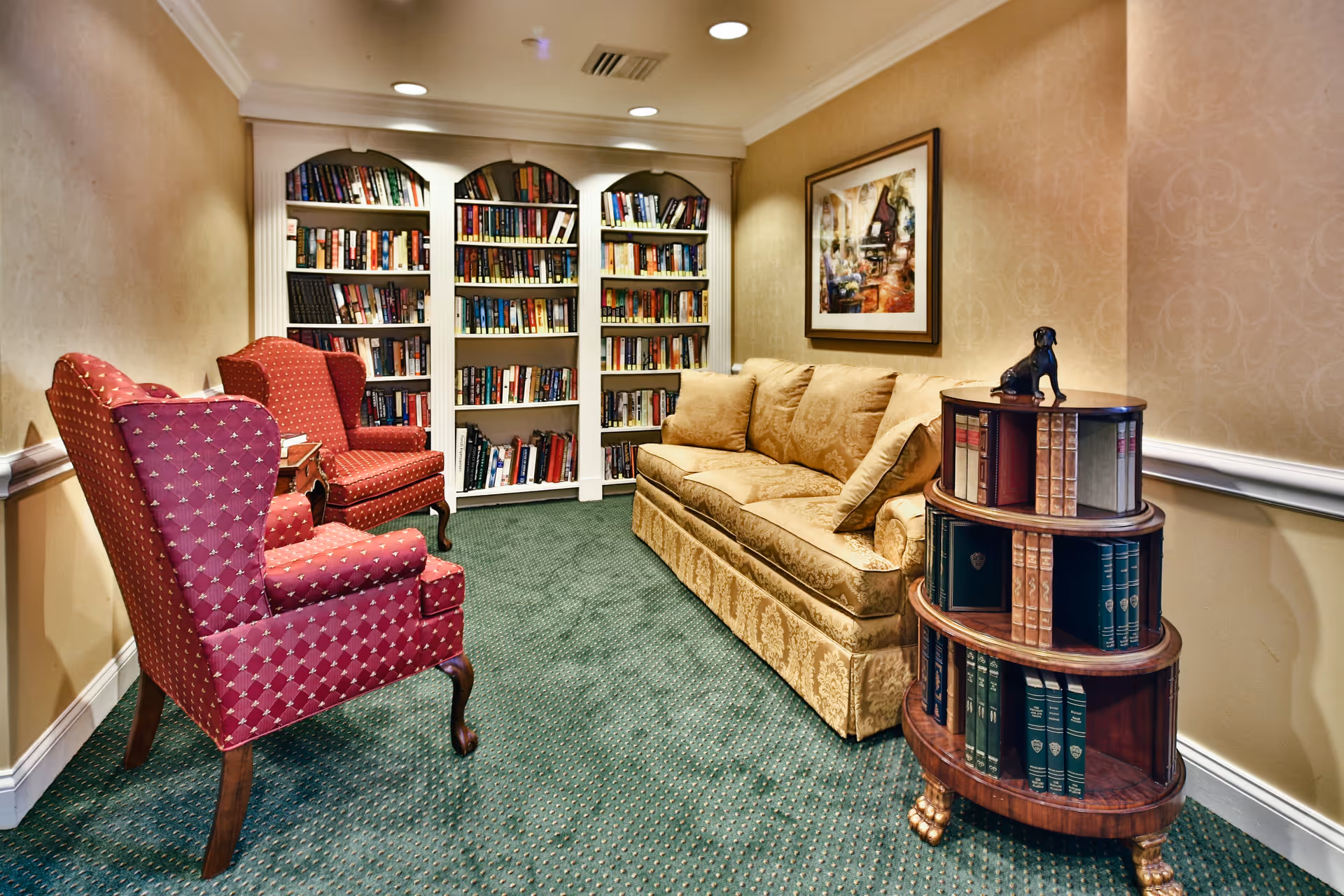 A cozy reading room with two red upholstered armchairs, a beige patterned sofa, a round wooden bookshelf filled with books, and a large white bookshelf against the wall. The room has green carpet, beige wallpaper, and a framed painting on the wall.