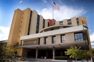 Exterior view of a multi-story nursing and rehabilitation center building with an American flag and another flag on flagpoles in front. The building has large windows and a covered entrance with trees and shrubs around.