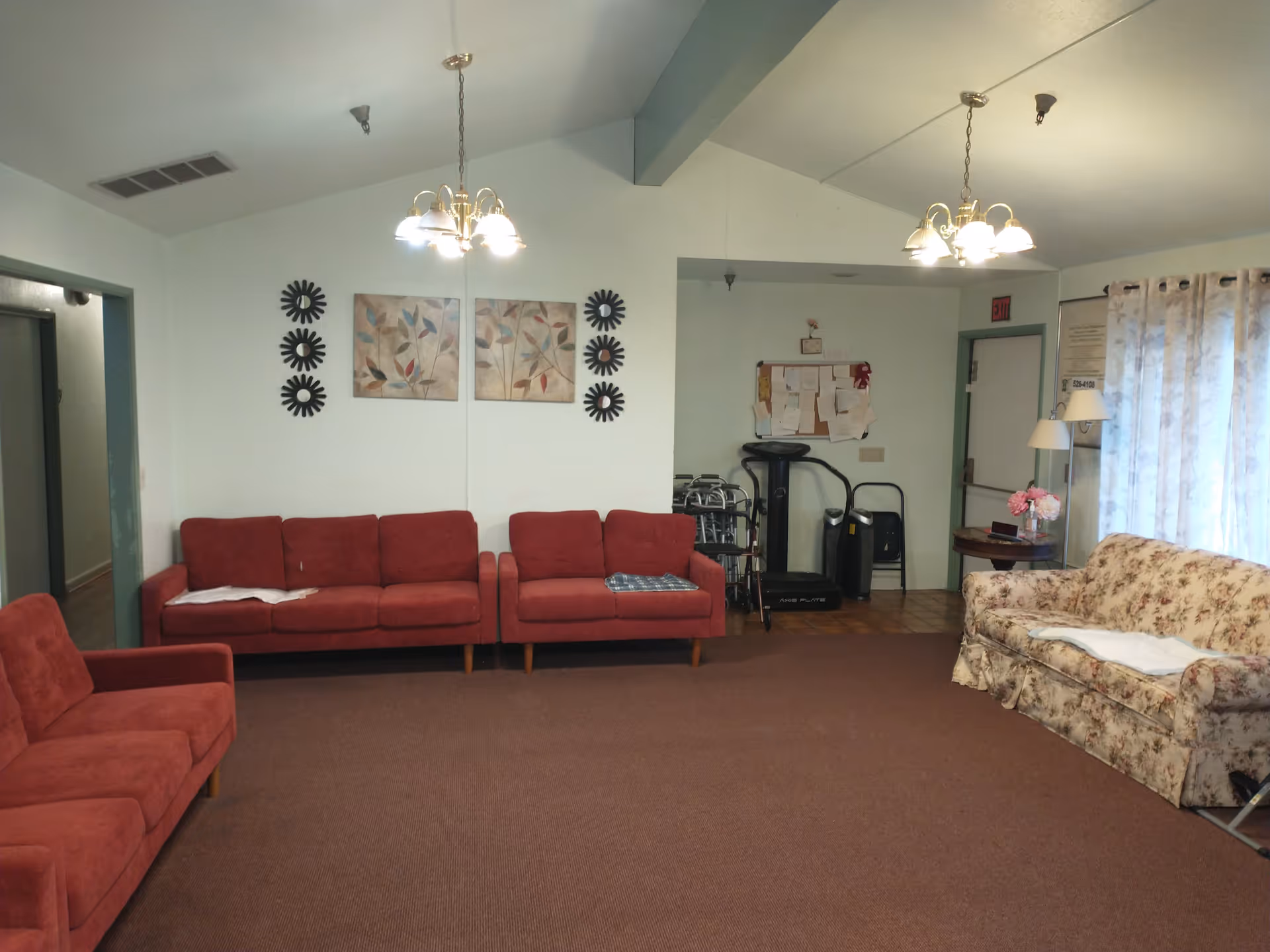 A senior living facility common area with red and floral patterned sofas arranged around the room. The walls are light-colored with decorative wall art and two ceiling light fixtures. There is a bulletin board with papers pinned on it, a walker, and some exercise equipment in the corner near a door with an exit sign above it. A floor lamp and a small round table with a flower vase are next to the floral sofa by a window with sheer curtains.