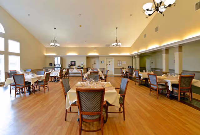 A spacious dining room with multiple tables covered in white tablecloths, each set with glassware and silverware. The room has wooden flooring, high ceilings with chandeliers, and large windows letting in natural light. The walls are painted in neutral tones with some green accents.