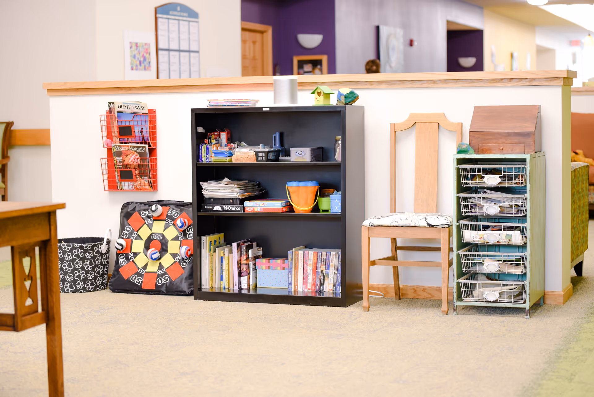Communal interior area with a bookshelf, chair, wire storage drawers, magazines and activity supplies against a half wall.
