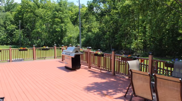 A spacious outdoor wooden deck with a grill and several chairs, surrounded by a railing with flower pots. The deck overlooks a grassy area and dense green trees in the background under a clear sky.