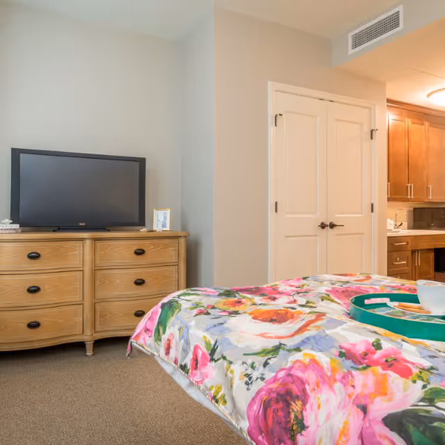 Interior view of a bedroom in Cogir of Old Town, Alexandria featuring a bed with a colorful floral bedspread, a wooden dresser with a flat-screen TV on top, and a kitchenette area with wooden cabinets in the background.