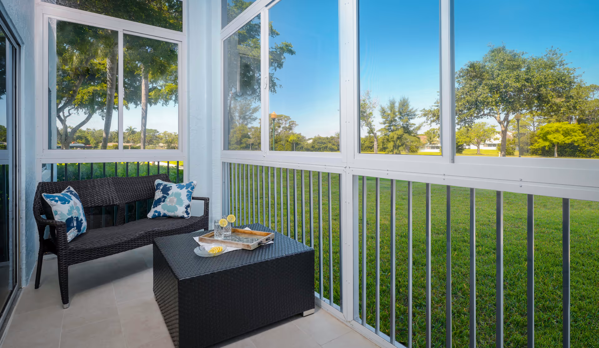 A screened-in porch with a black wicker loveseat adorned with two blue and white floral cushions, and a matching black wicker coffee table with a tray holding two glasses of lemon water and a small plate with lemon slices. The porch overlooks a green lawn with trees under a clear blue sky.