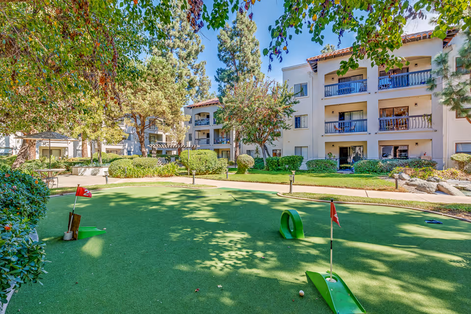 Outdoor putting green surrounded by trees and shrubs with a multi-story residential building in the background under a clear blue sky.