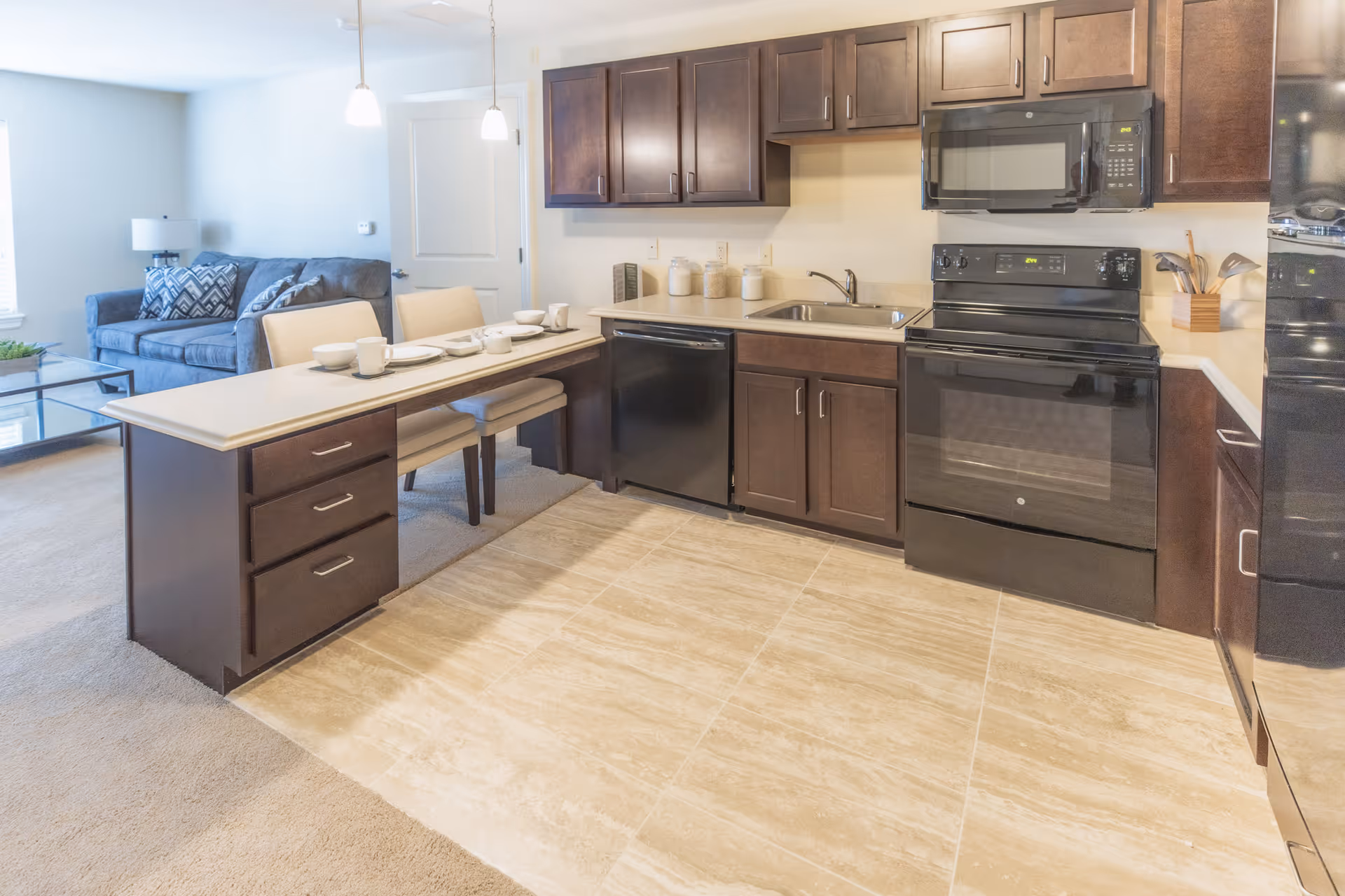 A modern kitchen with dark wood cabinets, black appliances including a microwave, stove, dishwasher, and refrigerator. There is a light-colored countertop with a sink and a peninsula with two chairs set with white dishes and cups. In the background, a living area with a gray couch, a lamp, and a glass coffee table is visible.
