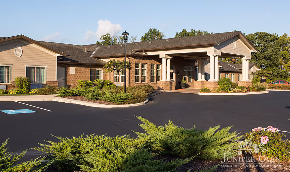 Exterior view of Juniper Glen Memory Care Assisted Living facility showing a single-story building with brick and beige siding, a covered entrance with white columns, a parking lot with a handicapped parking space, landscaped bushes, and trees under a clear sky.