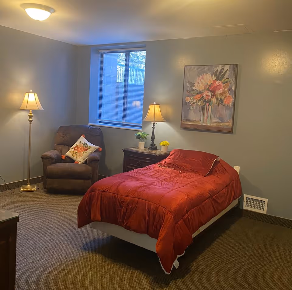 A cozy bedroom with a single bed covered in a red comforter, a wooden nightstand with two small potted plants and a lamp, a brown recliner chair with a floral pillow, a window with blinds, and a floral painting on the wall.