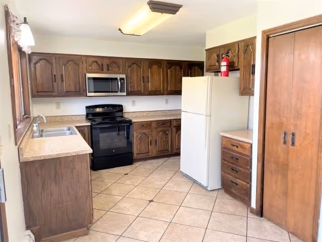 Kitchen with dark wood cabinets, tiled floor, white refrigerator, black stove and microwave, and a double sink.