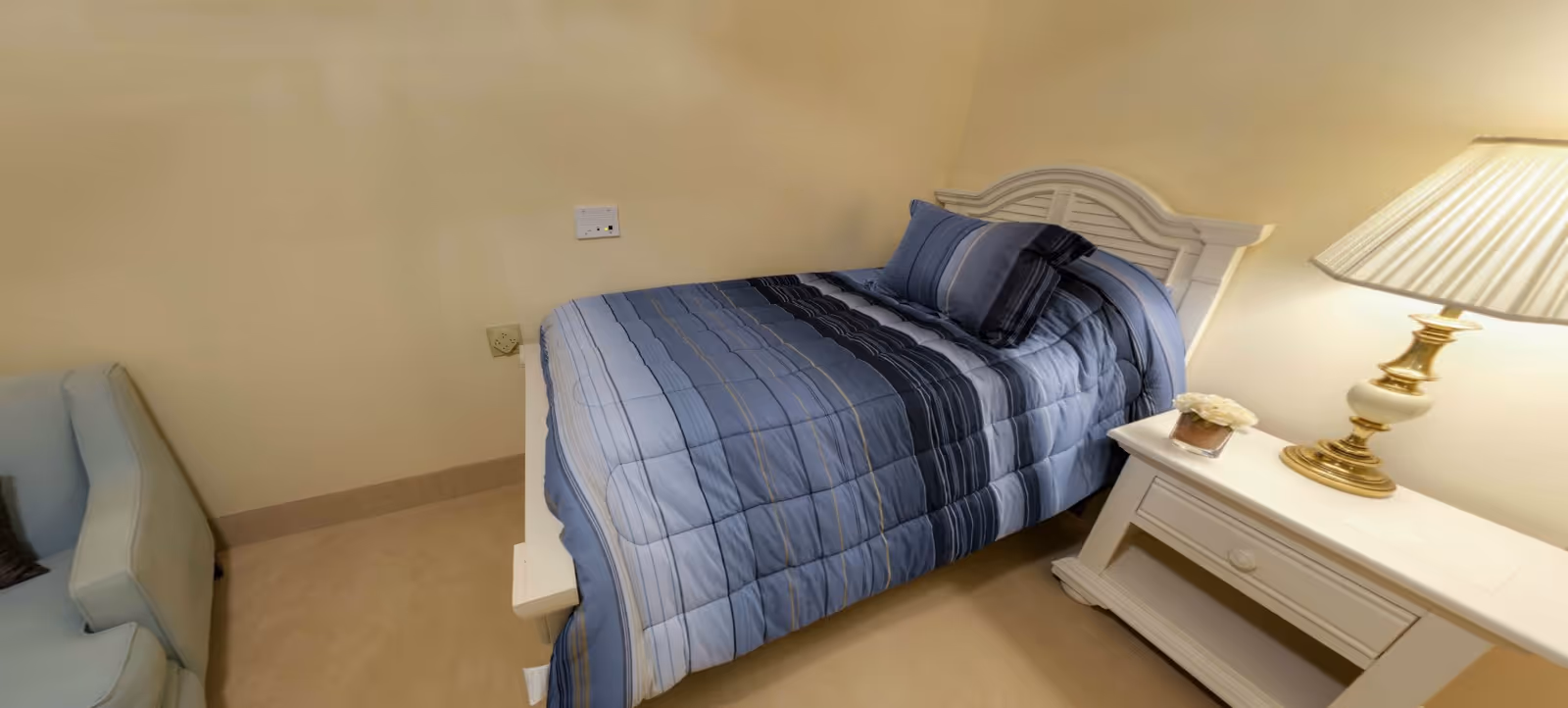 Small bedroom with a single bed dressed in blue striped bedding, a white nightstand with a lamp, and a chair.