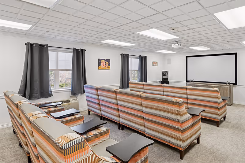 A senior living facility media room with rows of striped upholstered chairs facing a large projector screen. The room has white walls, two windows with dark curtains, a ceiling-mounted projector, and a popcorn machine in the corner.