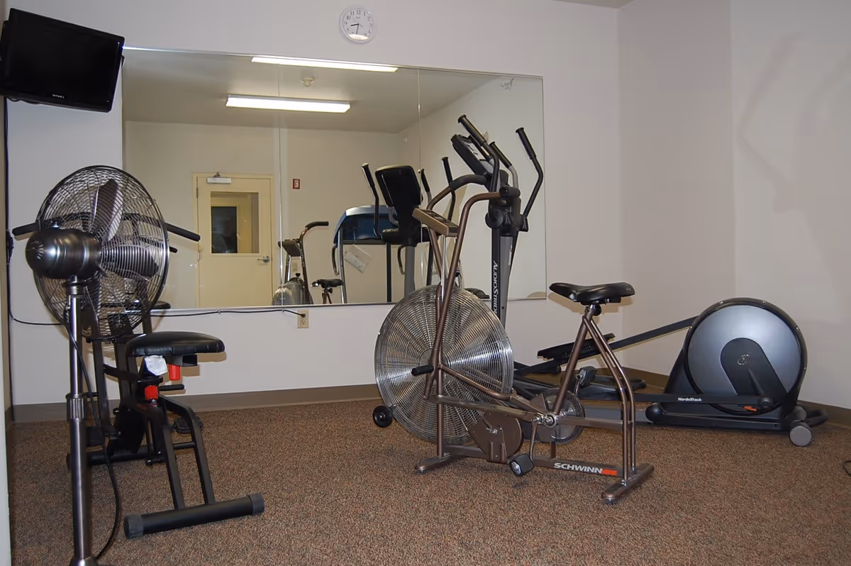 A small exercise room with various fitness equipment including a stationary bike, an elliptical machine, a rowing machine, and a large standing fan. A wall-mounted TV is visible in the upper left corner, and a large mirror covers most of the back wall reflecting the equipment and a closed door. A clock is mounted above the mirror.