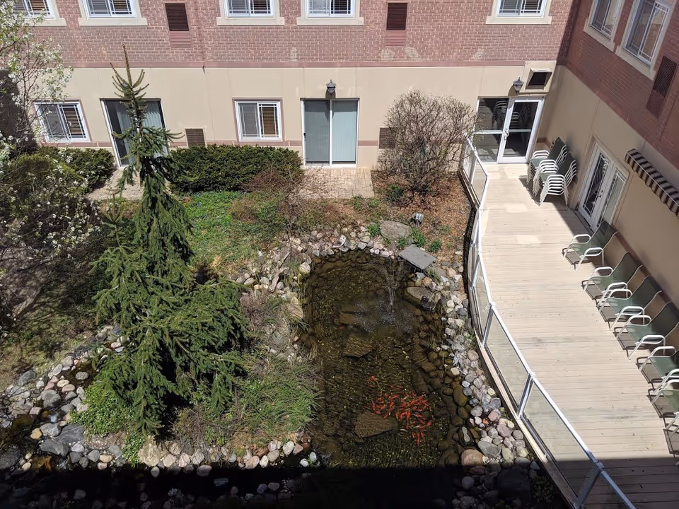 View of an outdoor courtyard at Franciscan Court Supportive Living featuring a small pond with fish, surrounded by rocks and greenery including a tall evergreen tree. There is a wooden deck with several chairs lined up along the building's exterior walls, and multiple windows and doors are visible on the building.