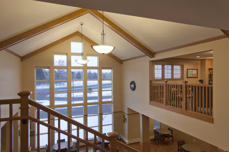 Interior view of a senior living facility with a high vaulted ceiling and large windows showing a snowy outdoor scene. The space includes a wooden staircase railing, a small seating area with tables and chairs on the lower level, and an upper balcony area with additional seating and bookshelves.