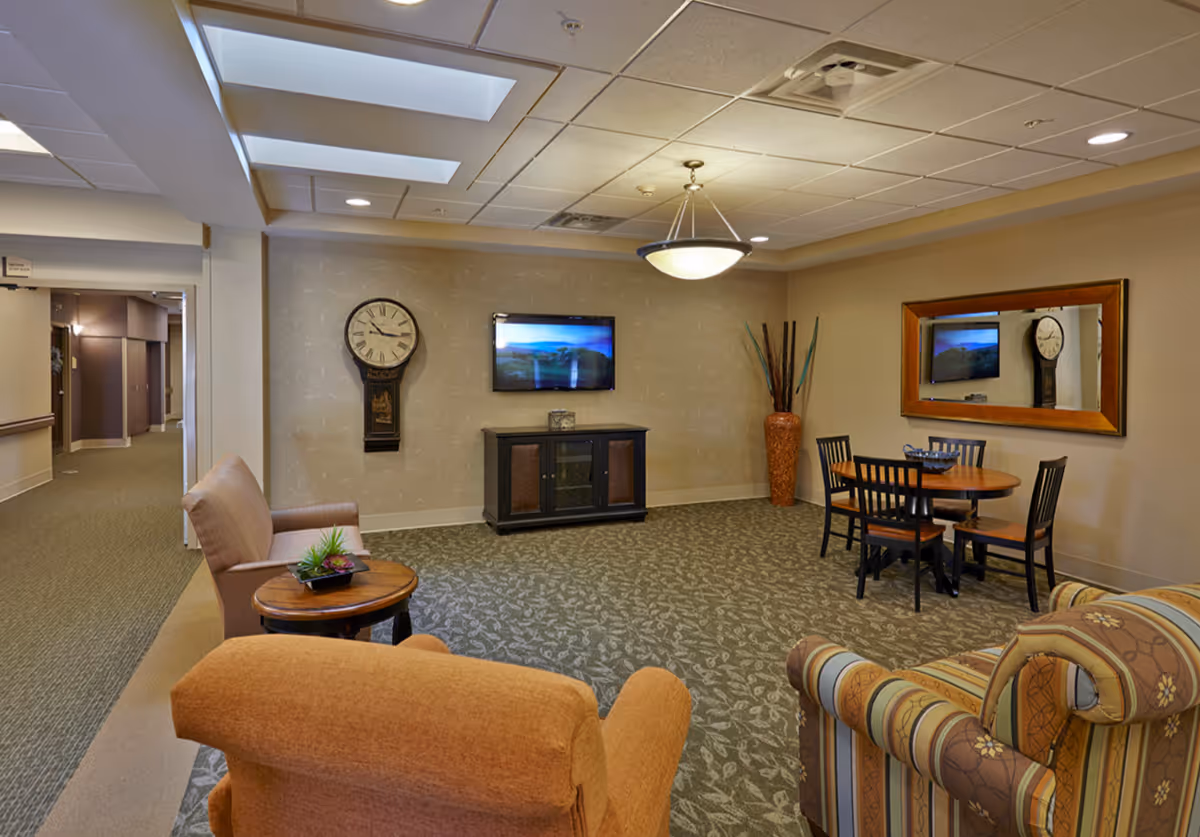 A cozy communal lounge with armchairs, a small dining table, a wall-mounted TV, decorative clock and mirror.