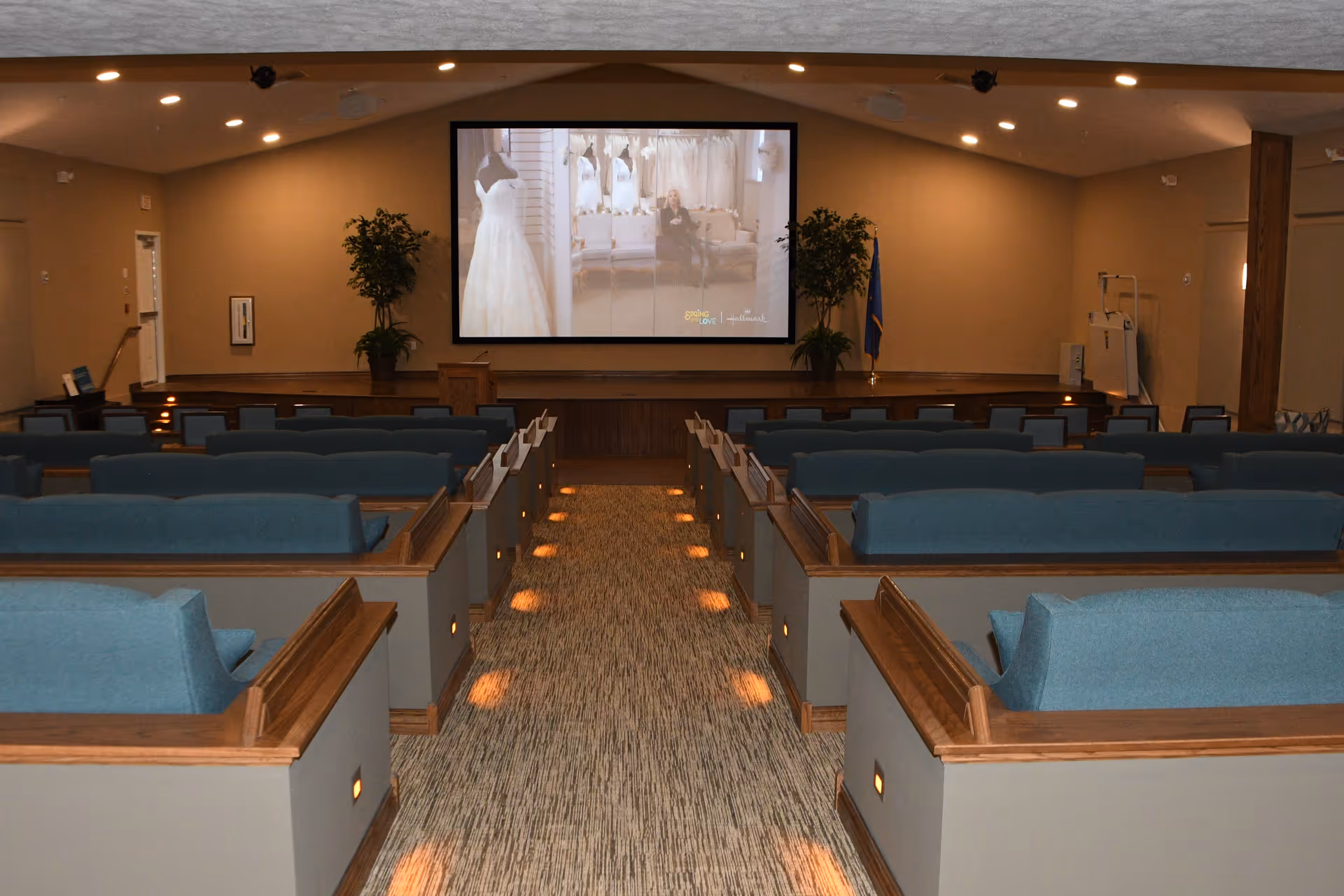 A small auditorium-style community room with rows of blue cushioned benches facing a large projection screen at the front.