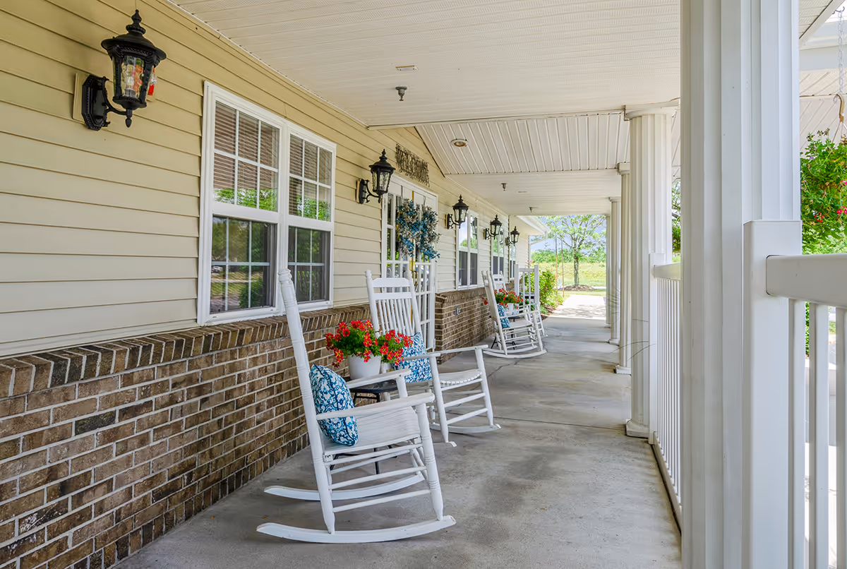 Covered porch area with white rocking chairs, each with a blue patterned cushion and a small table holding a pot of red flowers. The porch has beige siding with brick halfway up the wall, black wall-mounted lanterns, white columns, and a white railing. Greenery and trees are visible in the background.