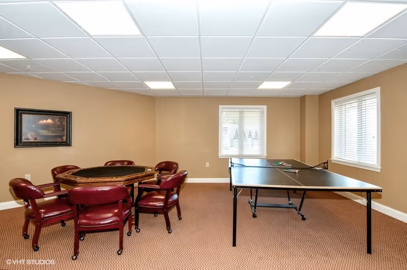 Carpeted game room with a poker table surrounded by red leather chairs and a ping-pong table near two windows.