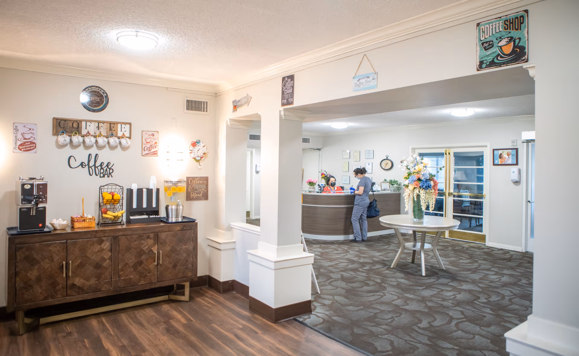 Interior view of a senior living facility lobby area with a coffee bar on the left featuring a coffee machine, cups, and fruit. Two people are interacting at a curved reception desk in the background. A round table with a large floral arrangement is in the center of the carpeted area. The walls are decorated with coffee-themed signs.