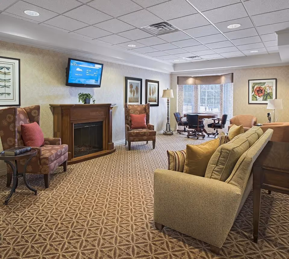 A cozy living room area in a senior living facility with patterned carpet, a beige sofa with multiple cushions, two patterned armchairs with red pillows, a wooden fireplace with a flat-screen TV mounted above it, framed artwork on the walls, a floor lamp, and a window with a table and chairs in the background.