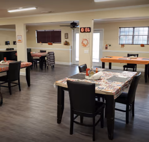 A communal dining room with several tables covered in patterned tablecloths and black chairs on wood-look flooring.