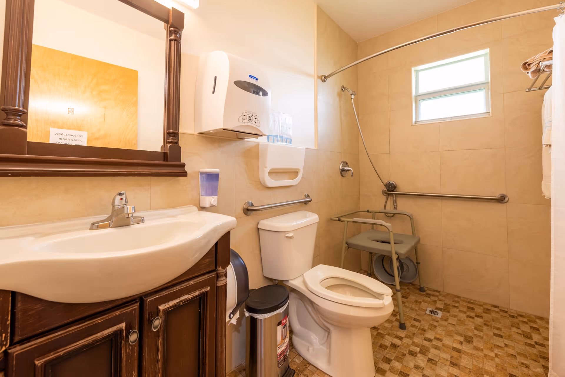 Bathroom with sink and mirror, toilet, grab bars, wall-mounted dispensers, and a shower area with a shower chair.