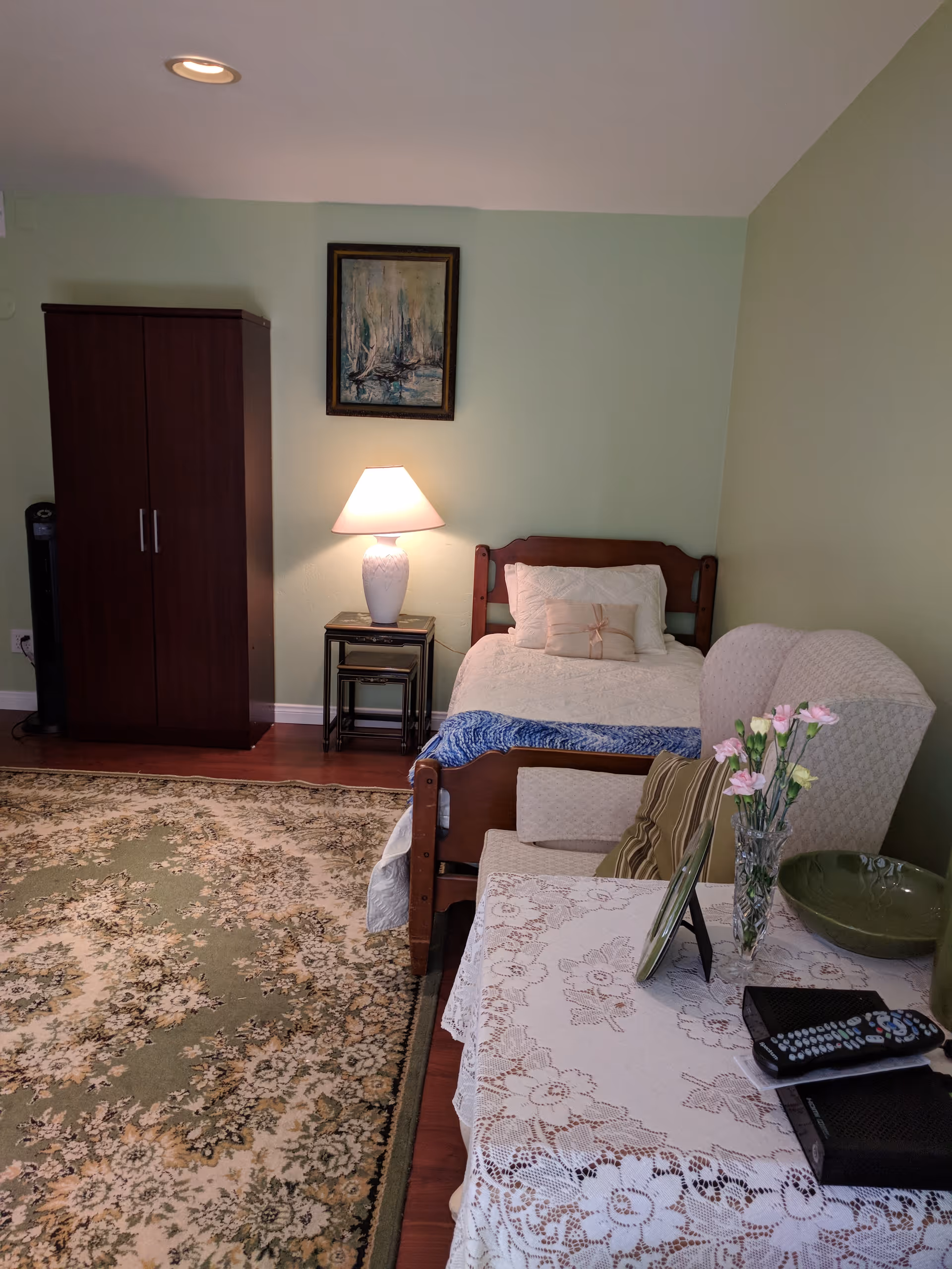 A cozy bedroom with a single wooden bed covered in white bedding and a blue throw. Next to the bed is a small table with a white lamp and a framed painting hanging above it. There is a dark wooden wardrobe against the wall and a patterned area rug covering the wooden floor. In the foreground, there is a table with a lace tablecloth, a vase with pink and yellow flowers, a green bowl, a remote control, and a small black device. A cushioned armchair with a striped pillow is partially visible beside the table.