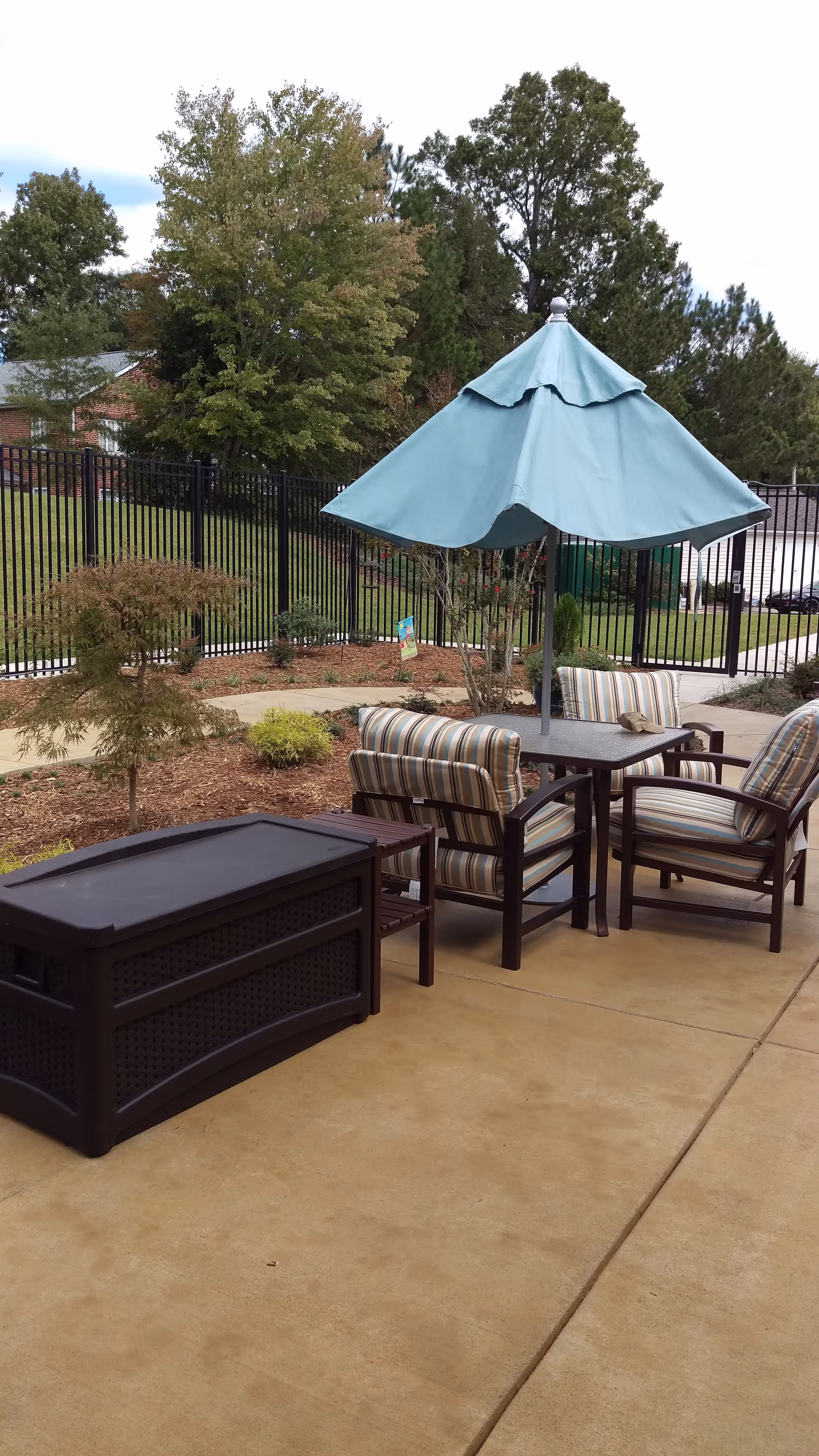 Outdoor patio area with a table, four cushioned chairs with striped fabric, and a large blue umbrella. The patio is surrounded by landscaping with small bushes and trees, and a black metal fence encloses the area.