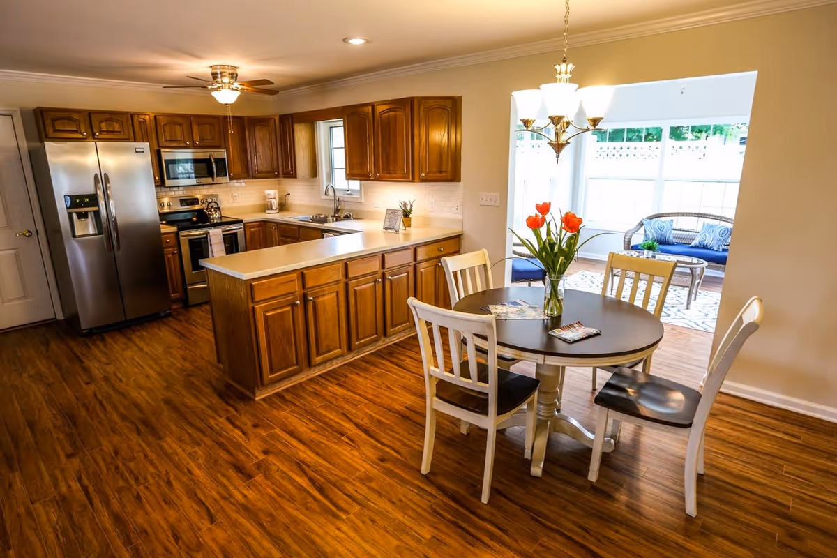 A modern kitchen featuring wooden cabinets, stainless steel appliances, and a round dining table with a vase of tulips. A sunroom with a blue couch is visible in the background.