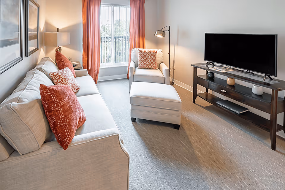 A bright living room with a beige sofa adorned with red and patterned throw pillows, a matching armchair with a red patterned pillow, and an ottoman. There is a floor lamp next to the armchair and a TV on a dark wooden stand with decorative items. The room has large windows with red curtains allowing natural light to enter.