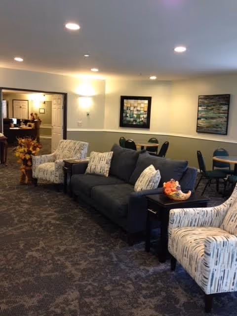 A cozy living room area in Holt Heritage Village featuring a dark gray sofa with patterned cushions, two patterned armchairs, side tables with decorative items, and round tables with chairs in the background. The walls are light-colored with two framed abstract paintings, and recessed ceiling lights illuminate the space.
