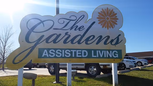 Outdoor sign reading "The Gardens Assisted Living" in front of a parking lot under a blue sky.