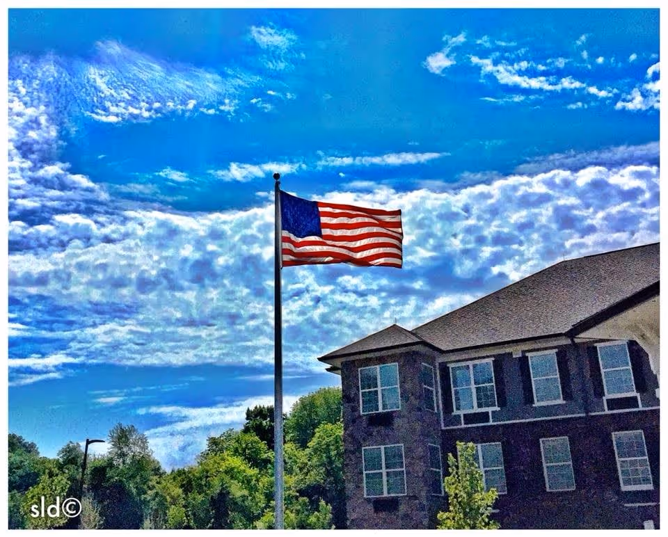 An American flag waving on a flagpole in front of a multi-story senior living facility building with multiple windows, under a partly cloudy blue sky.