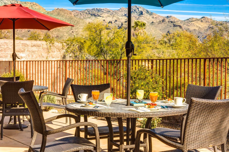 Outdoor patio area with round wicker tables and chairs under red and blue umbrellas. The tables have plates with food, glasses of juice, and coffee cups. In the background, there is a metal railing with desert vegetation and rocky mountains under a clear blue sky.