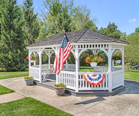 White wooden gazebo decorated with an American flag and red, white, and blue bunting, surrounded by green lawn and trees.