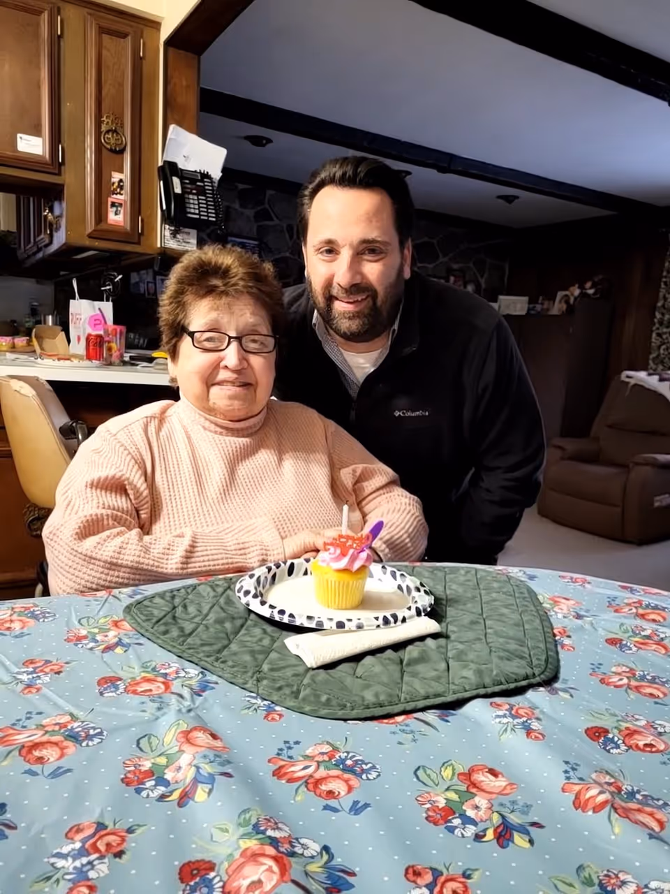 An elderly woman wearing glasses and a pink sweater sits at a table with a floral tablecloth and a green quilted placemat. In front of her is a plate with a yellow cupcake topped with pink frosting and a candle. A man with dark hair and a beard, wearing a black Columbia jacket, stands behind her smiling. The background shows a cozy interior with wooden cabinets, a phone, and a brown recliner chair.