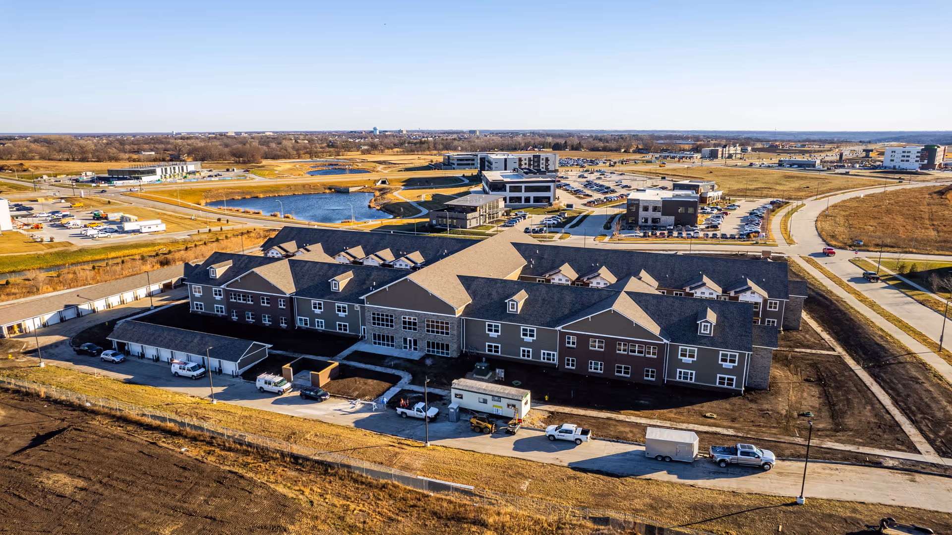 Aerial view of a large senior living facility building with a dark roof and multiple windows, surrounded by parking lots, roads, and open land with some ponds in the distance under a clear blue sky.