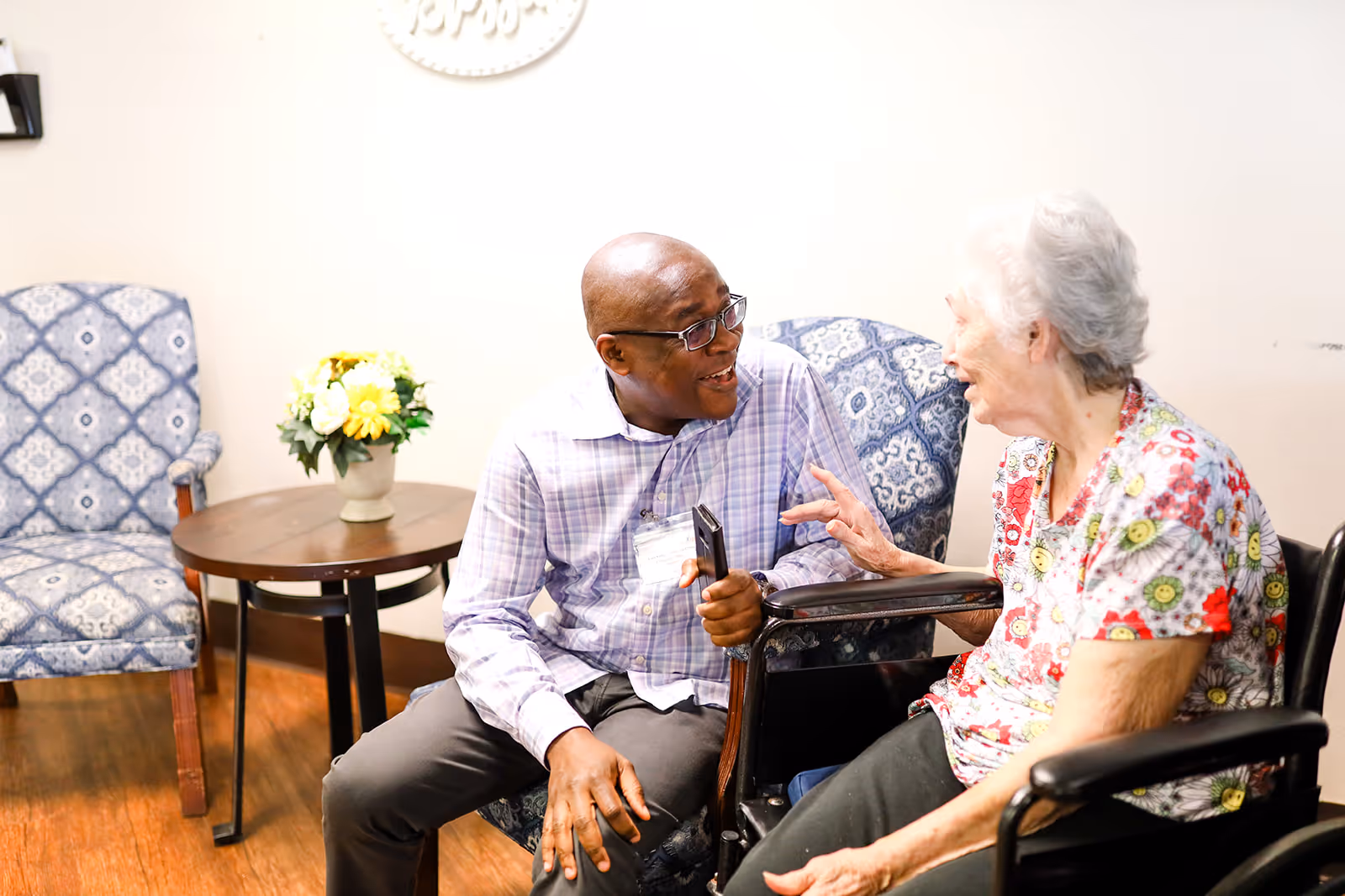 An elderly woman in a floral shirt sitting in a wheelchair is engaged in a lively conversation with a man wearing glasses and a plaid shirt, seated on a patterned armchair in a cozy room with wooden flooring and a small round table with a flower vase.