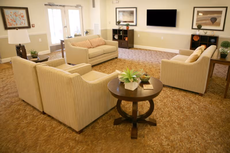A comfortable common room with beige sofas and armchairs arranged around a round wooden table with potted plants and a wall-mounted TV.