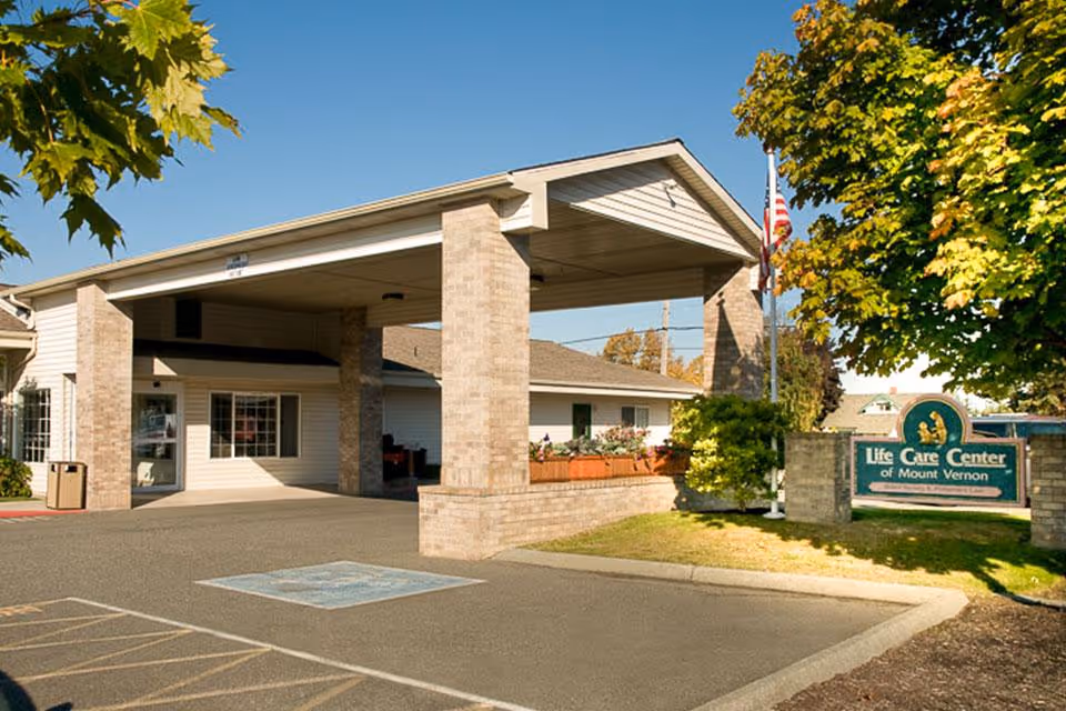 Exterior view of Life Care Center of Mount Vernon showing the entrance with a covered drop-off area supported by brick pillars, a parking area with handicap parking space, and a sign with the facility's name surrounded by trees and greenery under a clear blue sky.