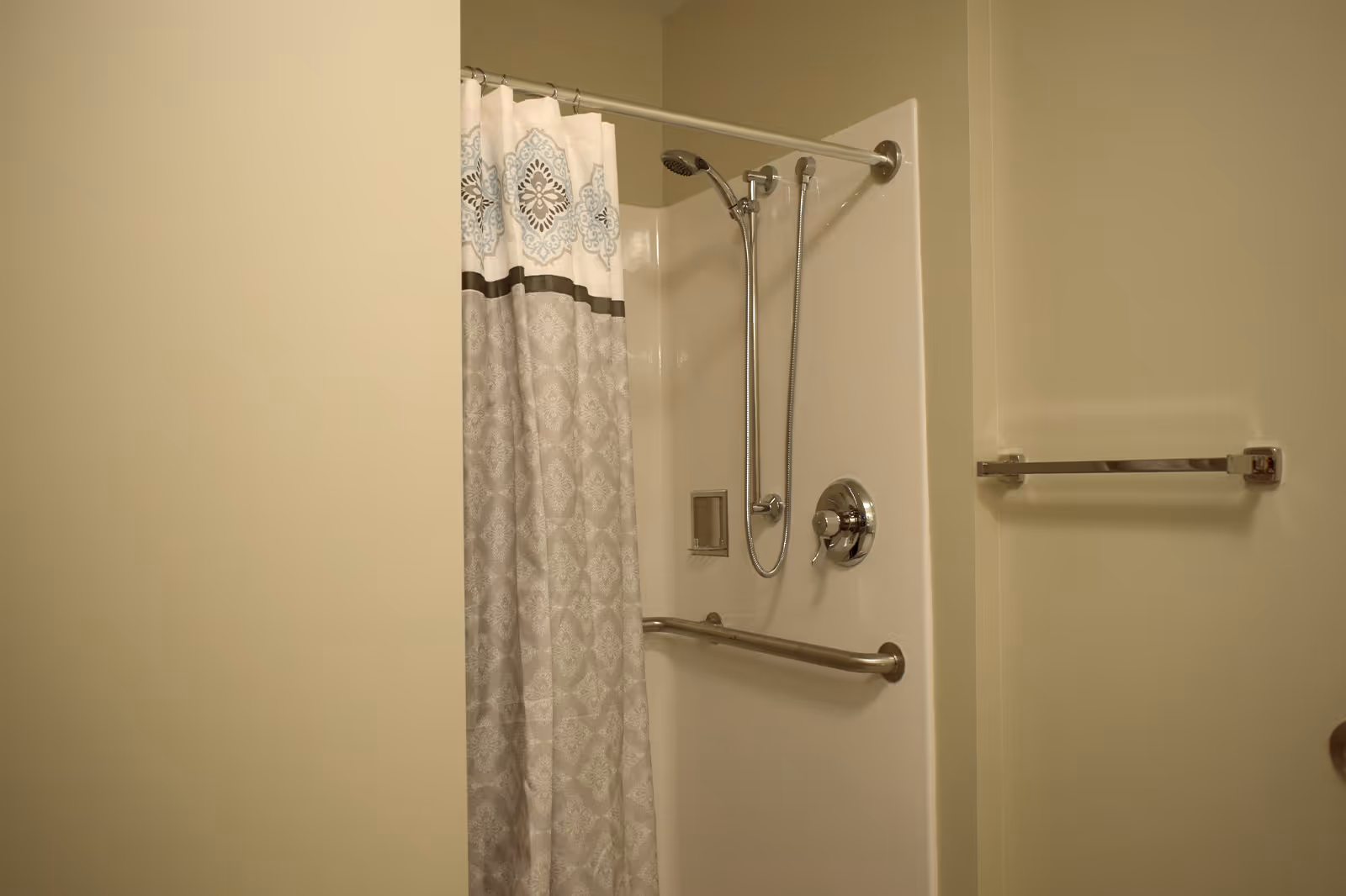 Shower stall with a patterned curtain, handheld showerhead, stainless grab bar and towel rack in a beige bathroom.