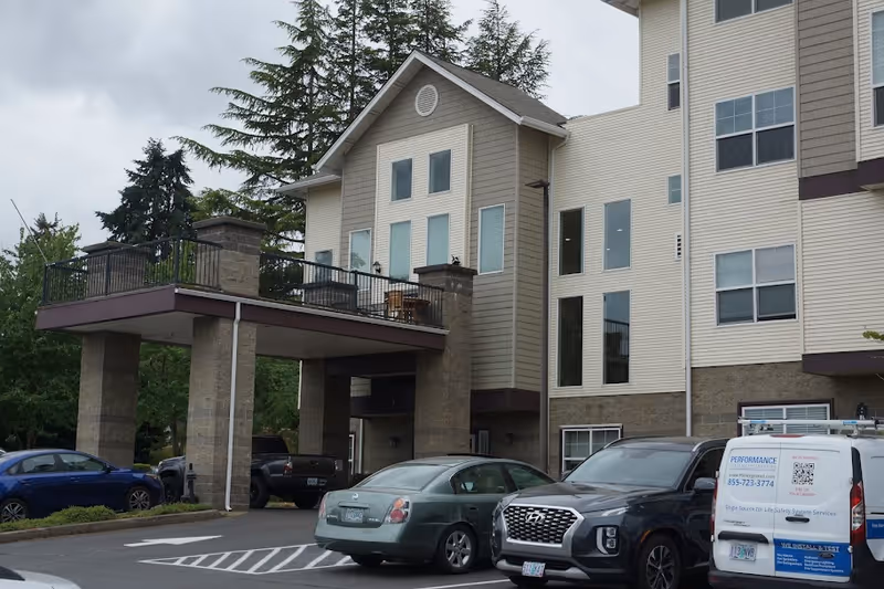 Exterior front entrance of a multi-story senior living building with a covered porte-cochere, balcony, and parked cars.