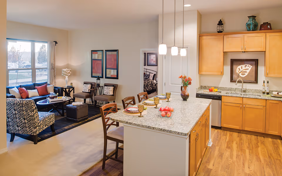 A bright and cozy open-concept living and kitchen area in a senior living facility. The living room features a large window with natural light, a patterned armchair, a dark coffee table, a sofa with colorful cushions, and two side chairs. The kitchen has wooden cabinets, granite countertops, a dishwasher, and a kitchen island with three chairs, set with plates, glasses, and a bowl of apples. Pendant lights hang above the island, and a bedroom is visible through an open door in the background.