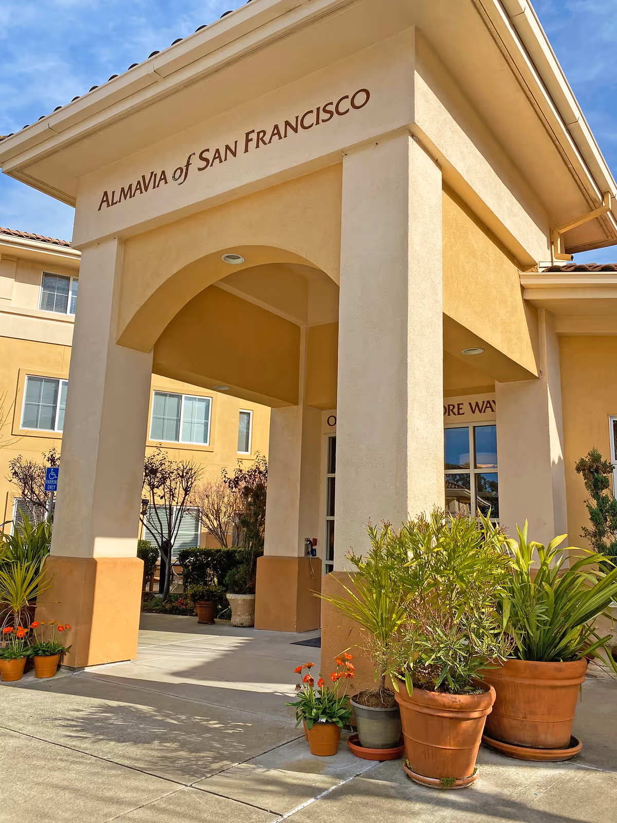 Entrance of AlmaVia of San Francisco facility with a covered archway supported by columns. Several potted plants and flowers are placed around the entrance area. The building exterior is beige with windows visible in the background under a partly cloudy sky.
