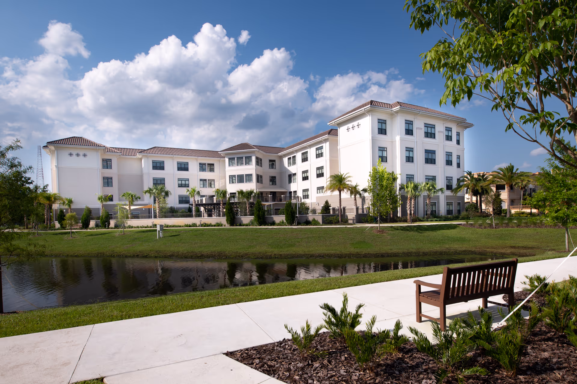 Exterior view of The Mayflower at Winter Park, a large multi-story senior living facility with white walls and many windows, surrounded by palm trees and greenery. A small pond and a bench on a paved walkway are visible in the foreground under a partly cloudy blue sky.