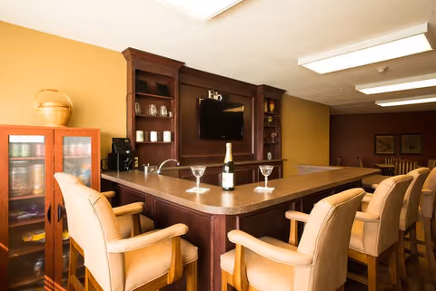 Interior view of a senior living facility lounge area with a bar counter surrounded by six cushioned bar stools. On the counter, there is a bottle of champagne and two empty cocktail glasses. Behind the bar, there is a mounted flat-screen TV and shelves with glassware. The room has warm lighting and a cozy atmosphere.
